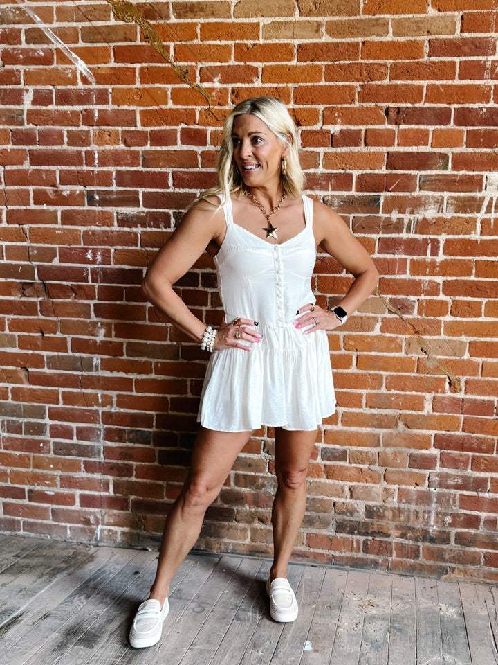 White romper with a sweetheart neckline and tiered skirt, worn by a woman standing against a brick wall.