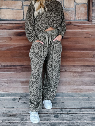 A person wearing a matching olive green and black leopard print lounge set, consisting of a long-sleeve top and wide-leg drawstring pants, standing on a rustic wooden deck in front of a stone wall.