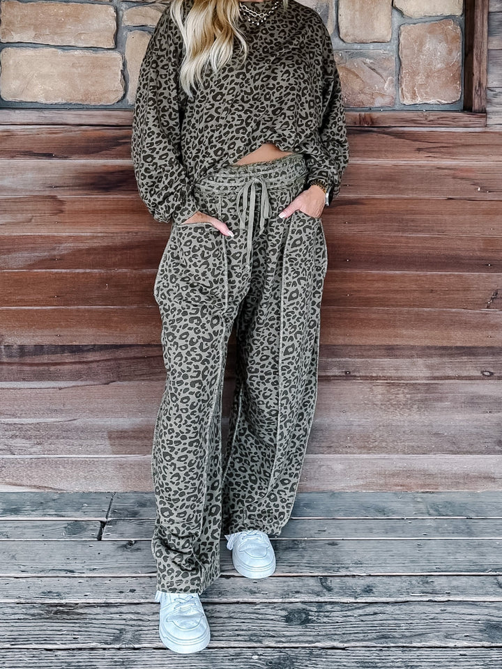 A person wearing a matching olive green and black leopard print lounge set, consisting of a long-sleeve top and wide-leg drawstring pants, standing on a rustic wooden deck in front of a stone wall.