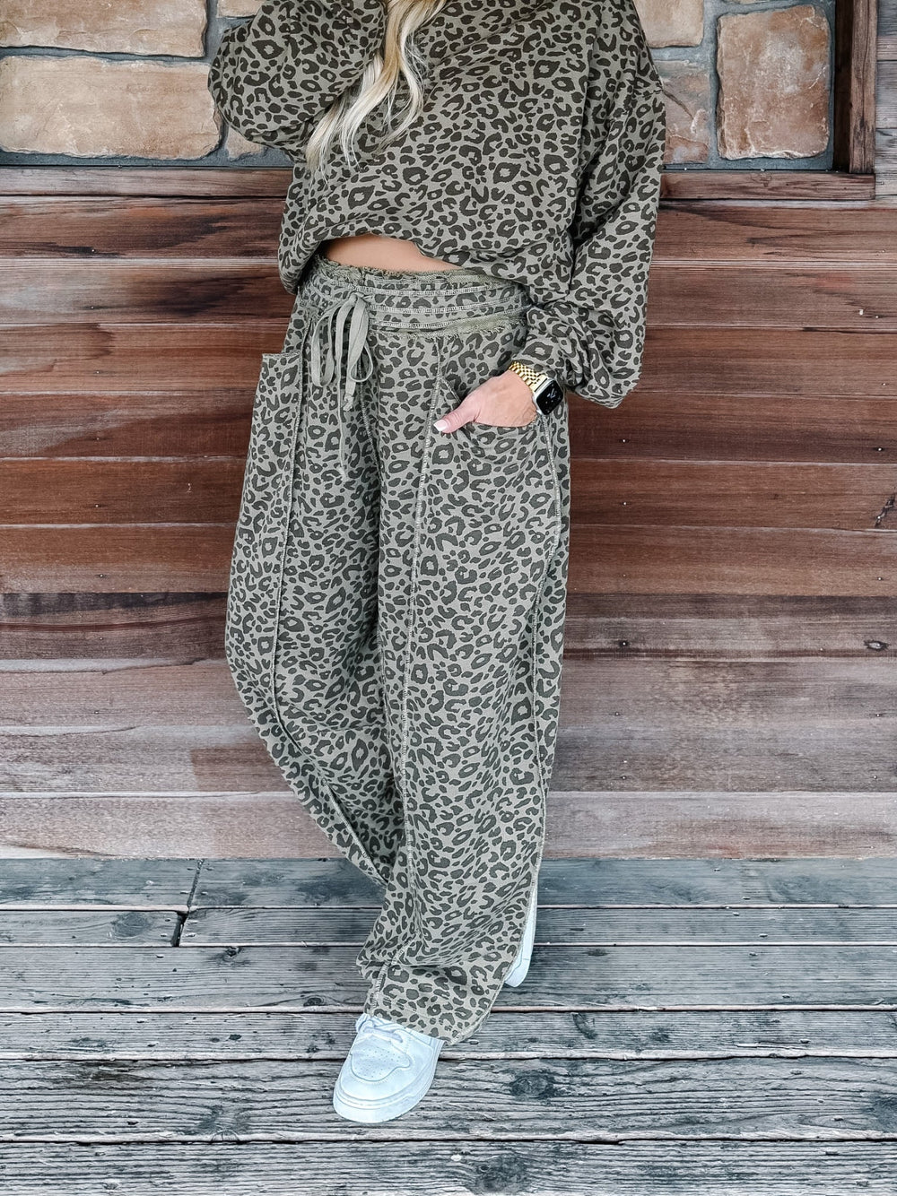 A person wearing a matching leopard-print lounge set with a long-sleeve top and wide-leg drawstring pants, standing on a weathered wooden deck in front of a wood plank wall.