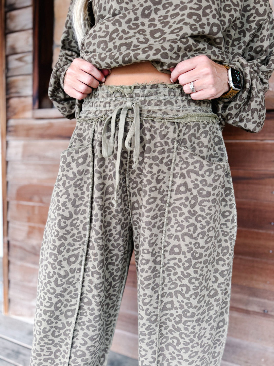 A person wearing loose leopard-print sweatpants with a drawstring waist and a matching sweatshirt, standing in front of a wooden wall.