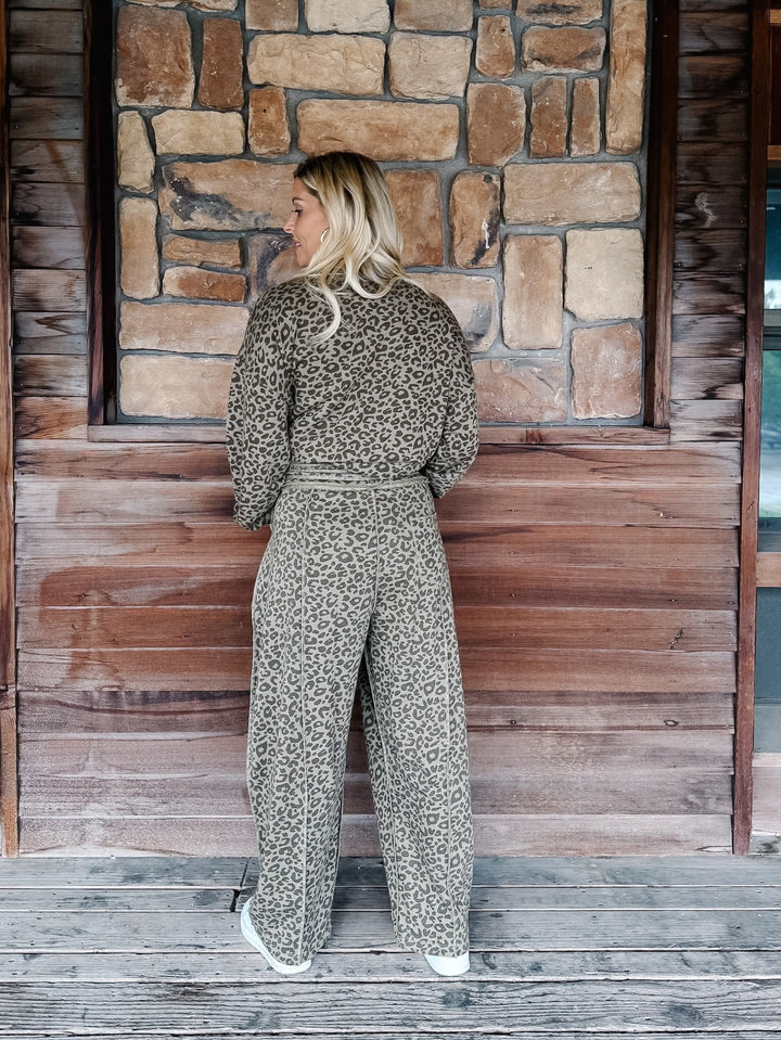 A woman stands on a rustic wooden porch with a stone wall background, wearing a matching beige and black leopard-print long-sleeve top and wide-leg pants set.