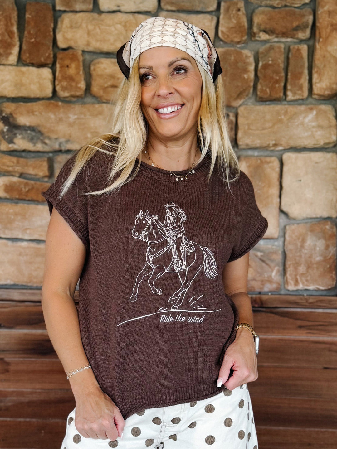 A smiling blonde woman wearing a brown short-sleeve knit t-shirt with a white line art graphic of a cowboy riding a horse and the text &