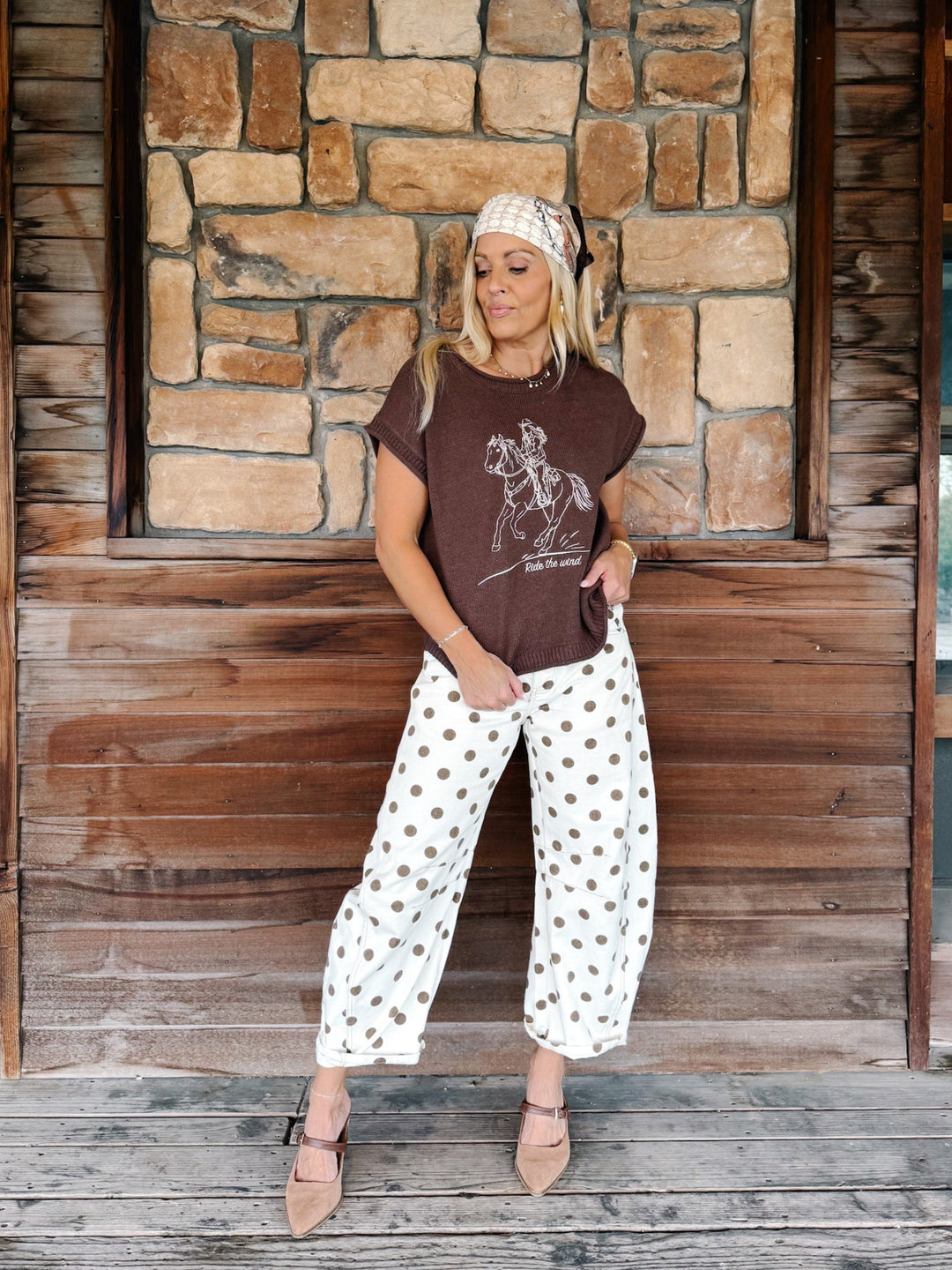 A woman stands on a wooden porch in front of a stone wall, wearing a brown graphic t-shirt, white cropped pants with brown polka dots, a beige headband, and tan heeled sandals.
