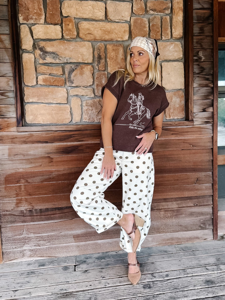 A woman posing on a rustic wooden porch in front of a stone wall, wearing a brown graphic t-shirt, white pants with brown polka dots, and a beige headband.
