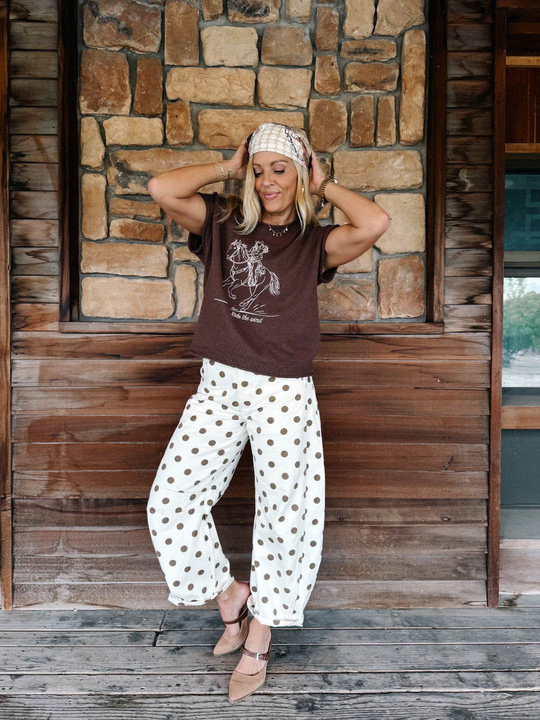A woman posing on a wooden porch in front of a stone wall, wearing a brown graphic t-shirt, white pants with brown polka dots, a patterned headband, and tan flat shoes.