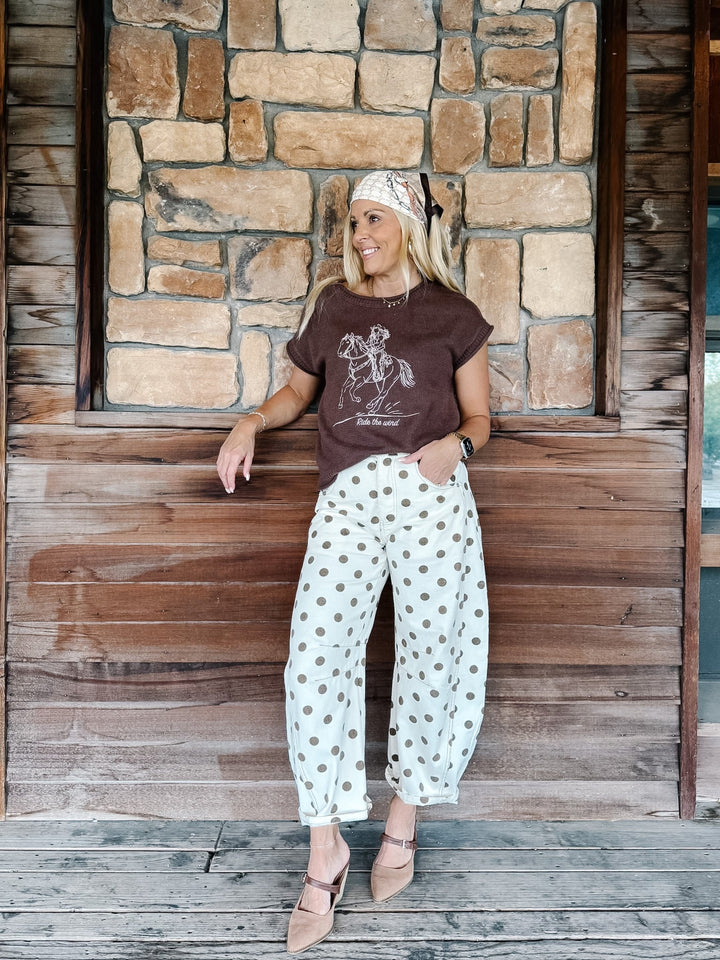 A woman posing on a rustic wooden porch in front of a stone wall, wearing a brown graphic t-shirt, white pants with brown polka dots, and beige flat shoes.