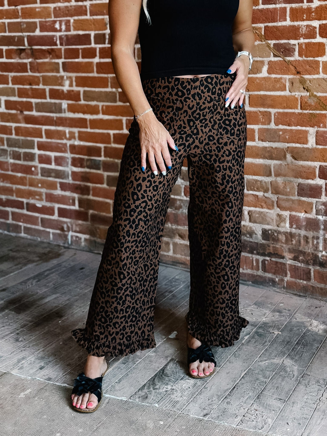 A person wearing loose, wide-leg leopard-print pants with a black top and black slide sandals, standing against a red-brown brick wall and wood floor.