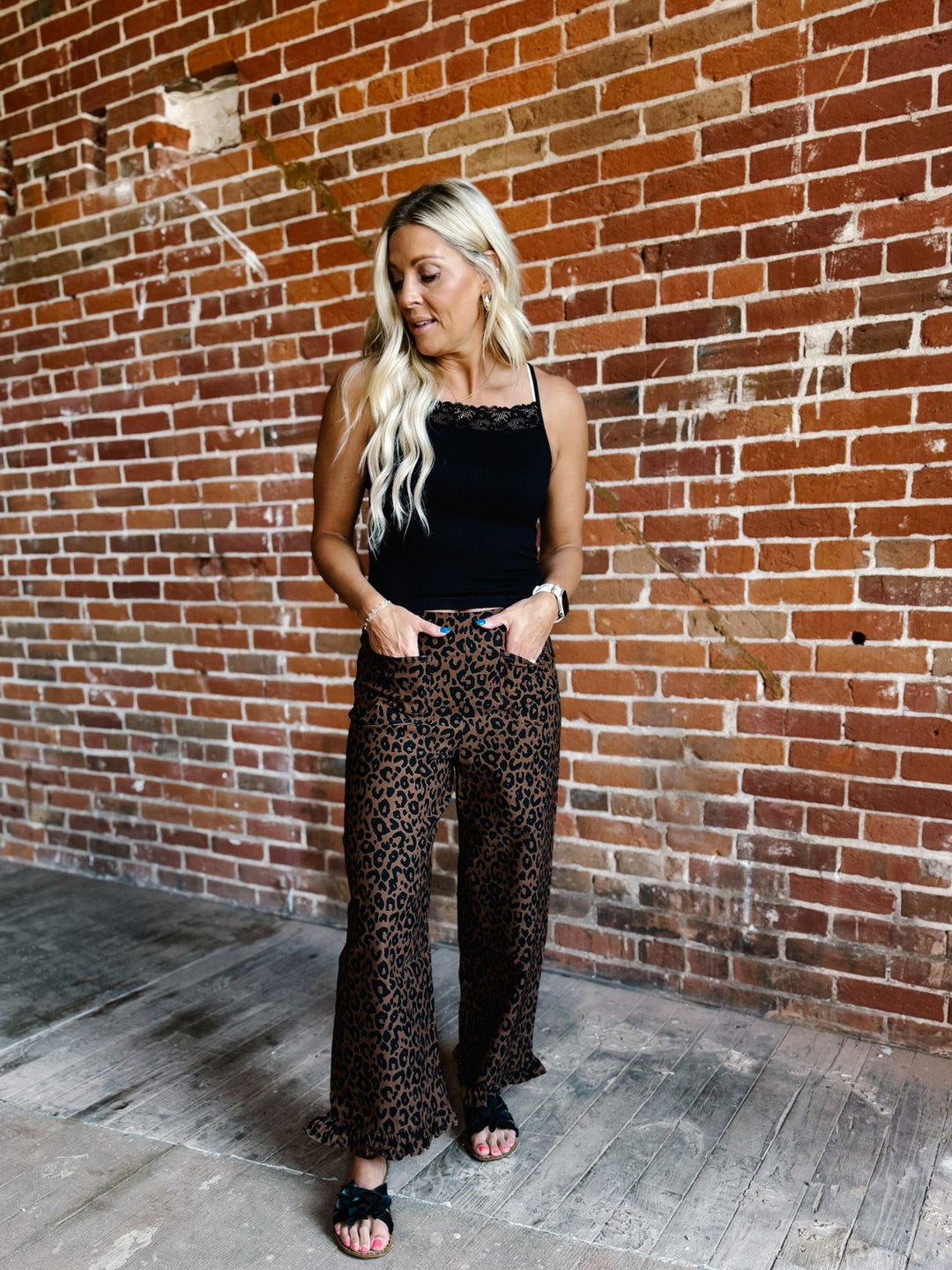 A woman stands against a red brick wall wearing leopard-print wide-leg pants and a black sleeveless top.