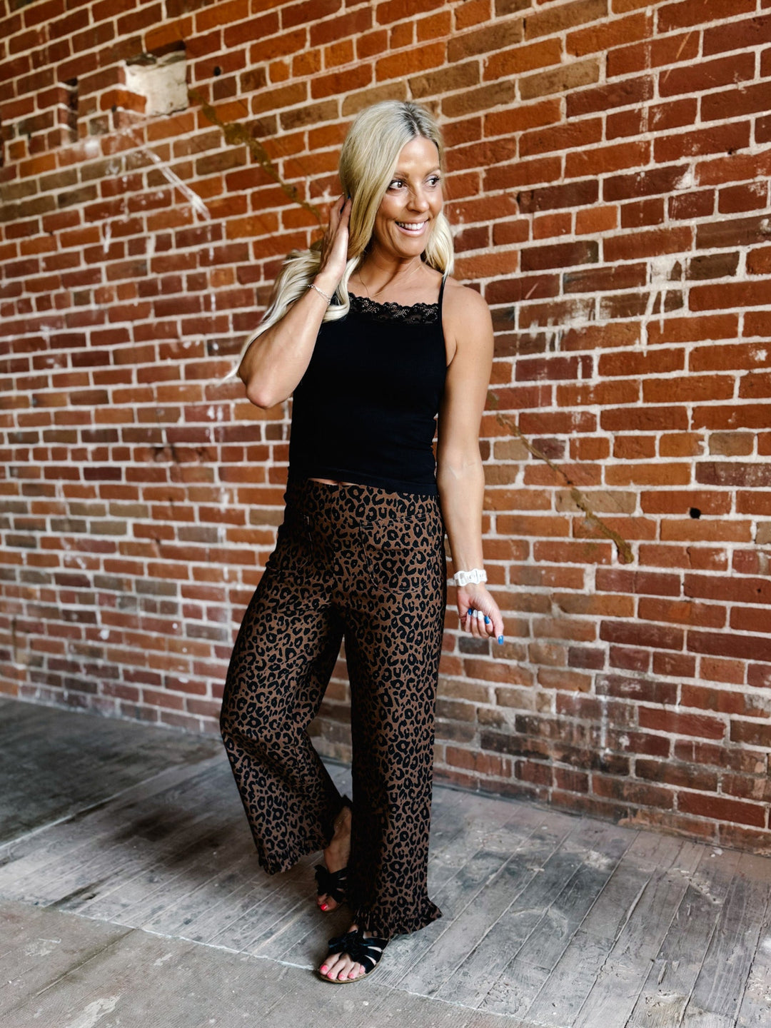 A woman posing in front of a brick wall wearing a black lace-trimmed camisole top and leopard-print wide-leg pants.