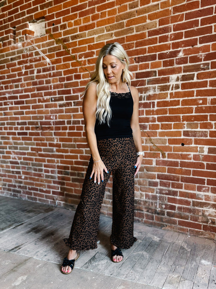 A woman posing in front of a red brick wall, wearing a black sleeveless top and black wide-leg pants with a brown and black leopard print, paired with black slide sandals.