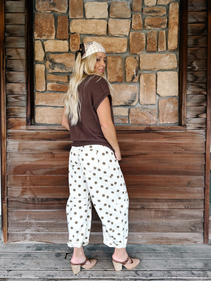 A woman stands on a wooden porch in front of a stone wall, wearing loose white pants with dark brown polka dots, a dark brown short-sleeve top, a white headband, and beige heeled sandals.