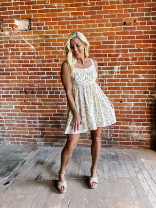 A woman wearing a floral mini dress with a scoop neckline and adjustable tie back, standing against a brick wall.