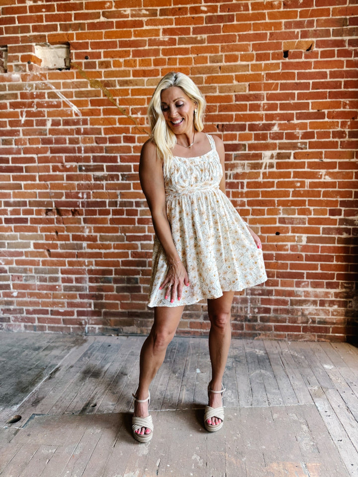 A woman wearing a floral mini dress with a scoop neckline and adjustable tie back, standing against a brick wall.