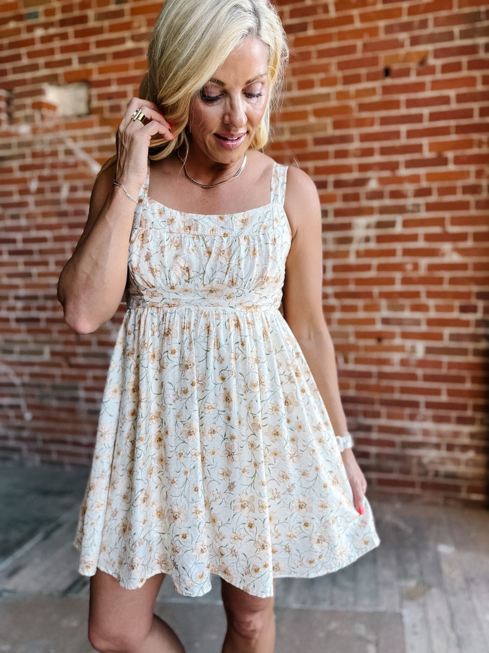 A woman wearing a floral mini dress with a scoop neckline and adjustable tie back, standing against a brick wall.