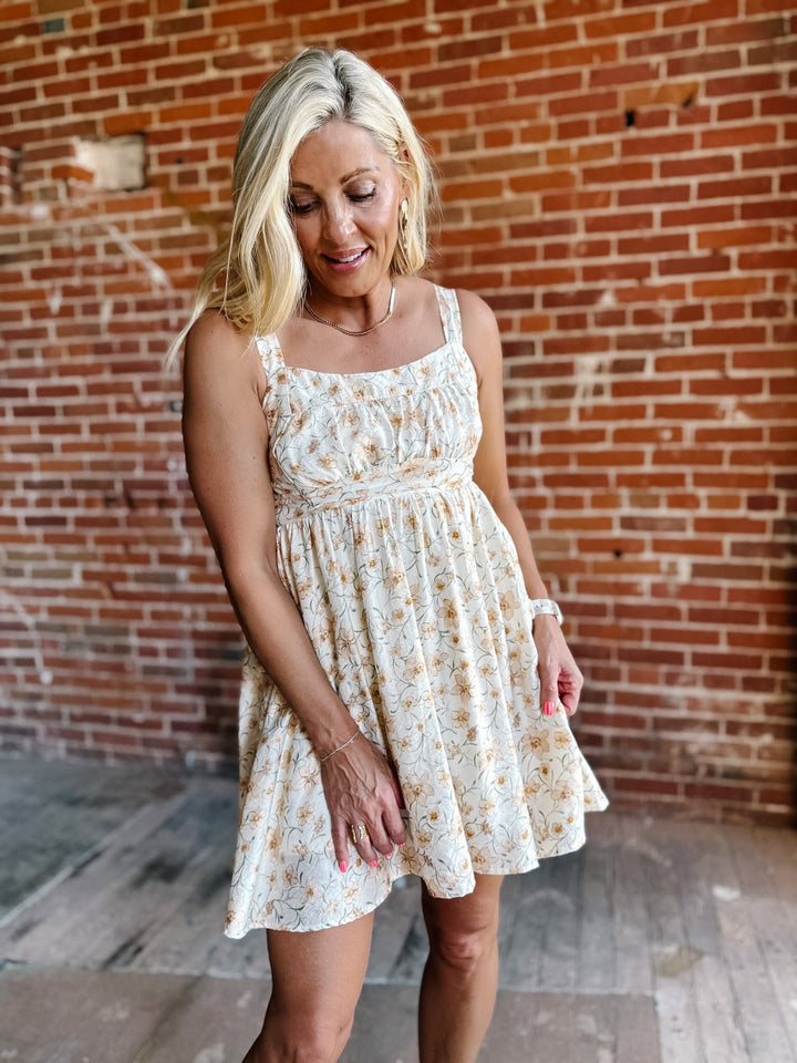 A woman wearing a floral mini dress with a scoop neckline and adjustable tie back, standing against a brick wall.