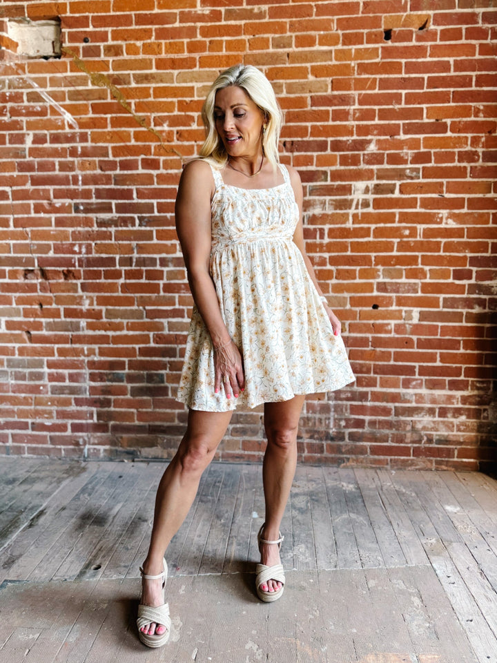 A woman wearing a floral mini dress with a scoop neckline and adjustable tie back, standing against a brick wall.