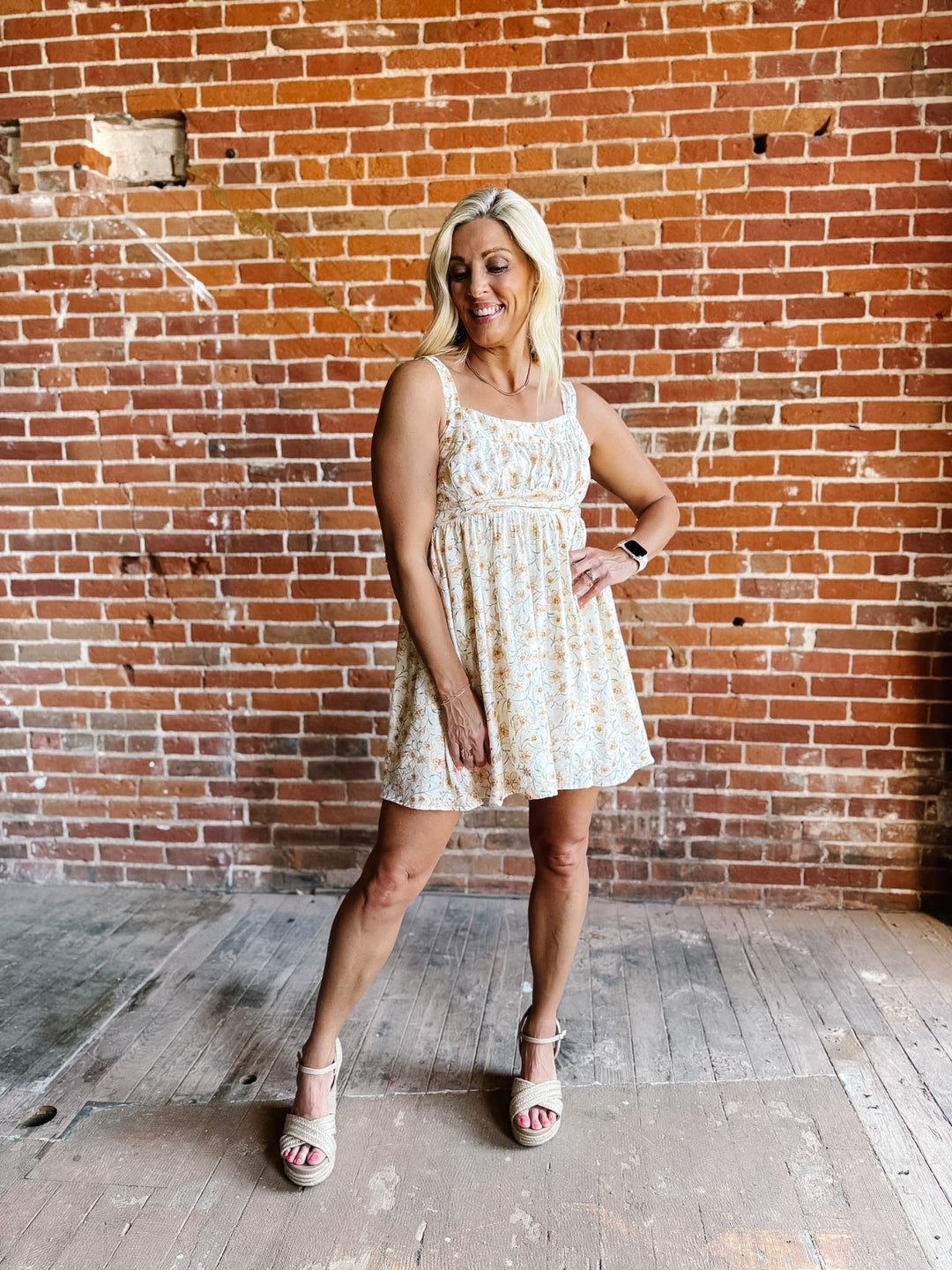 A woman wearing a floral mini dress with a scoop neckline and adjustable tie back, standing against a brick wall.