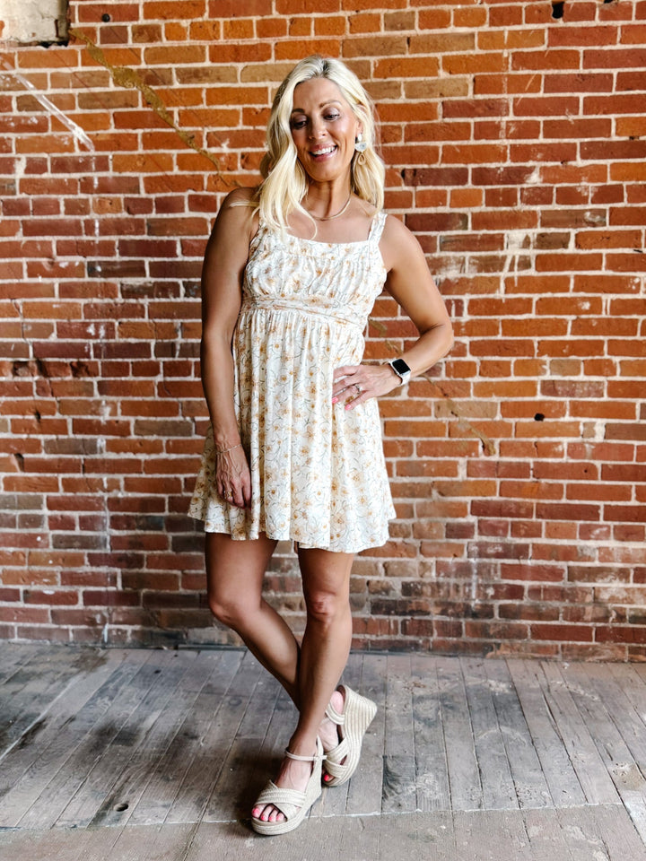 A woman wearing a floral mini dress with a scoop neckline and adjustable tie back, standing against a brick wall.