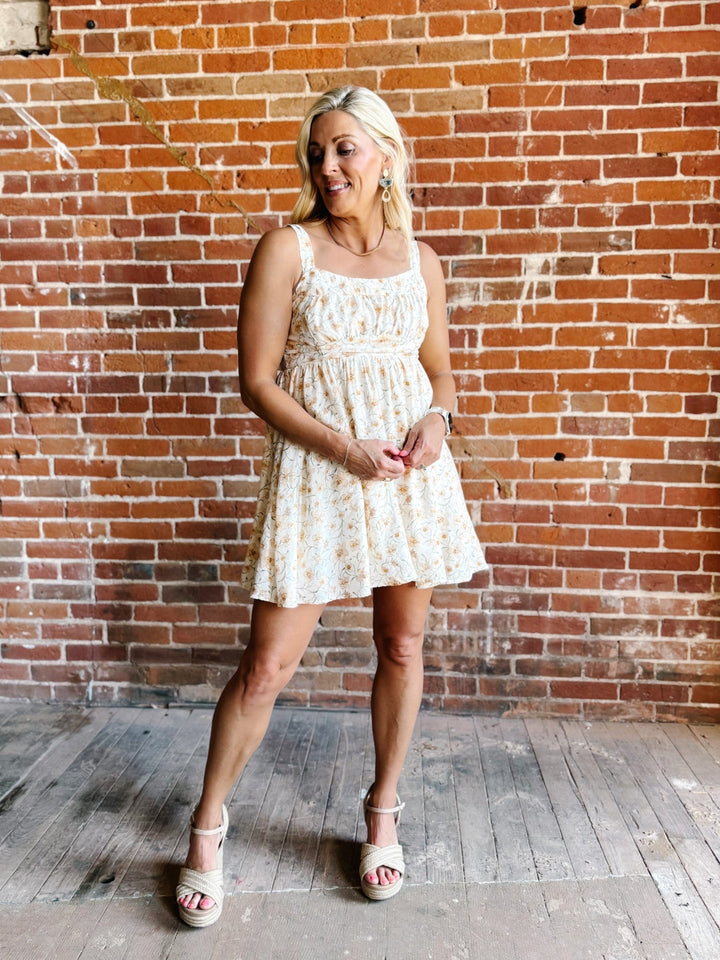 A woman wearing a floral mini dress with a scoop neckline and adjustable tie back, standing against a brick wall.
