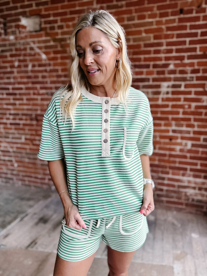 A woman wearing a green zig zag stripe lounge set with a Henley top and shorts, standing against a brick wall.