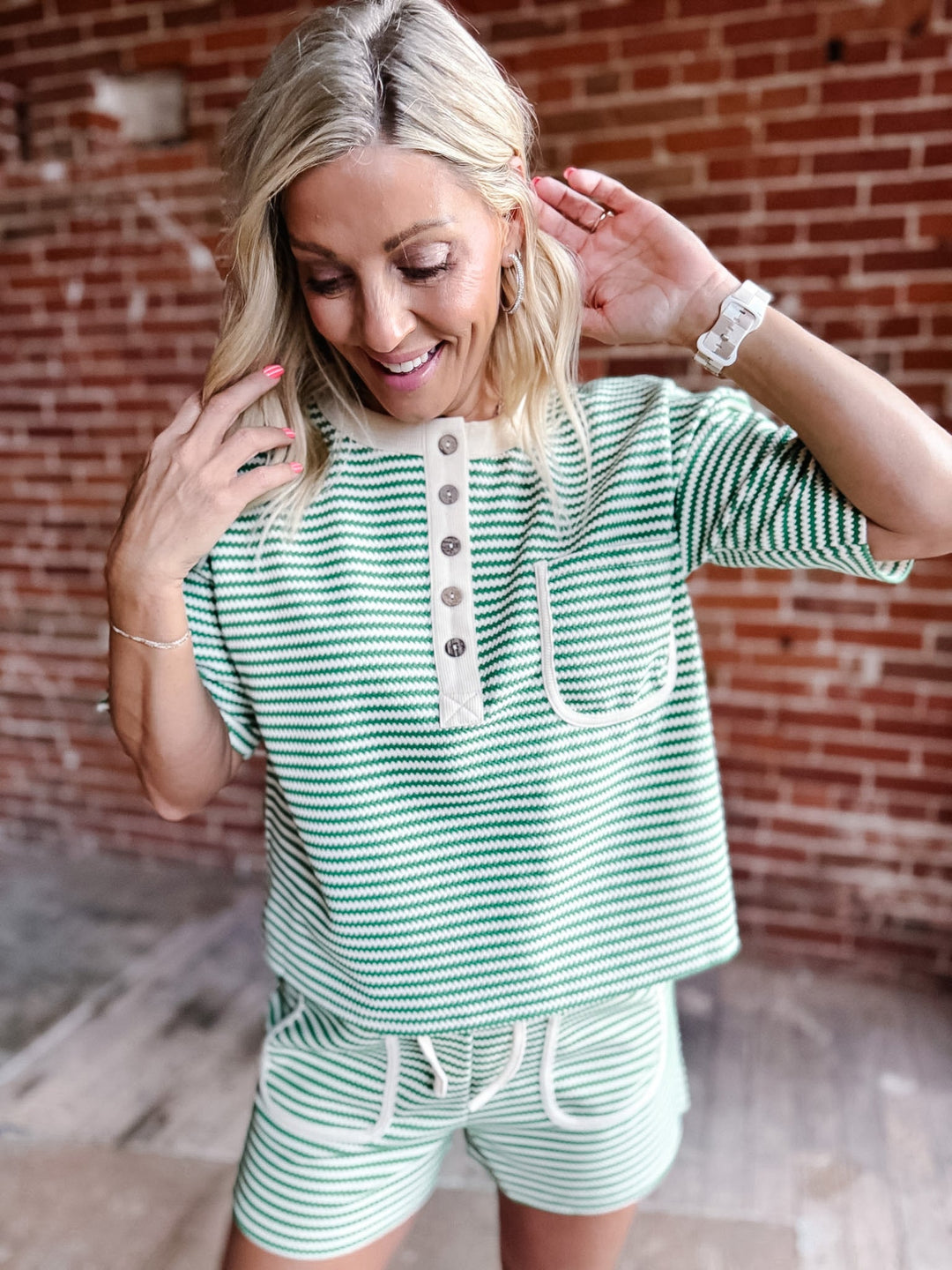 A woman wearing a green zig zag stripe lounge set with a Henley top and shorts, standing against a brick wall.