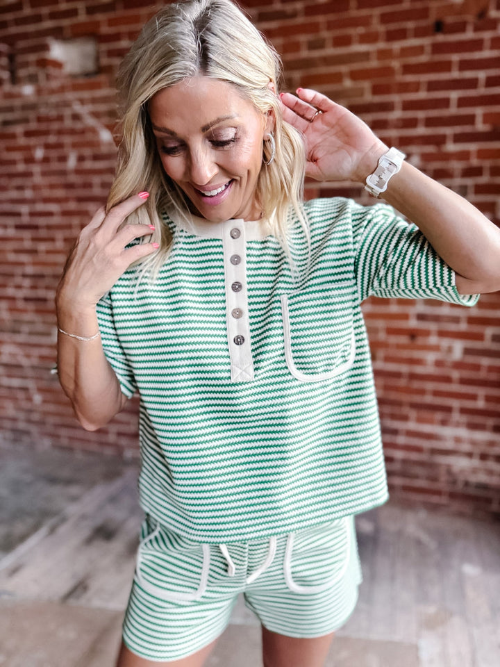 A woman wearing a green zig zag stripe lounge set with a Henley top and shorts, standing against a brick wall.