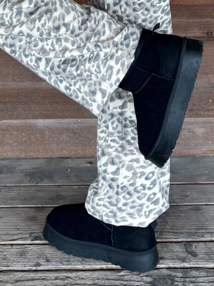 Black suede Chelsea-style slipper boots with chunky black soles, worn with white and gray leopard print pants on a weathered wooden deck.