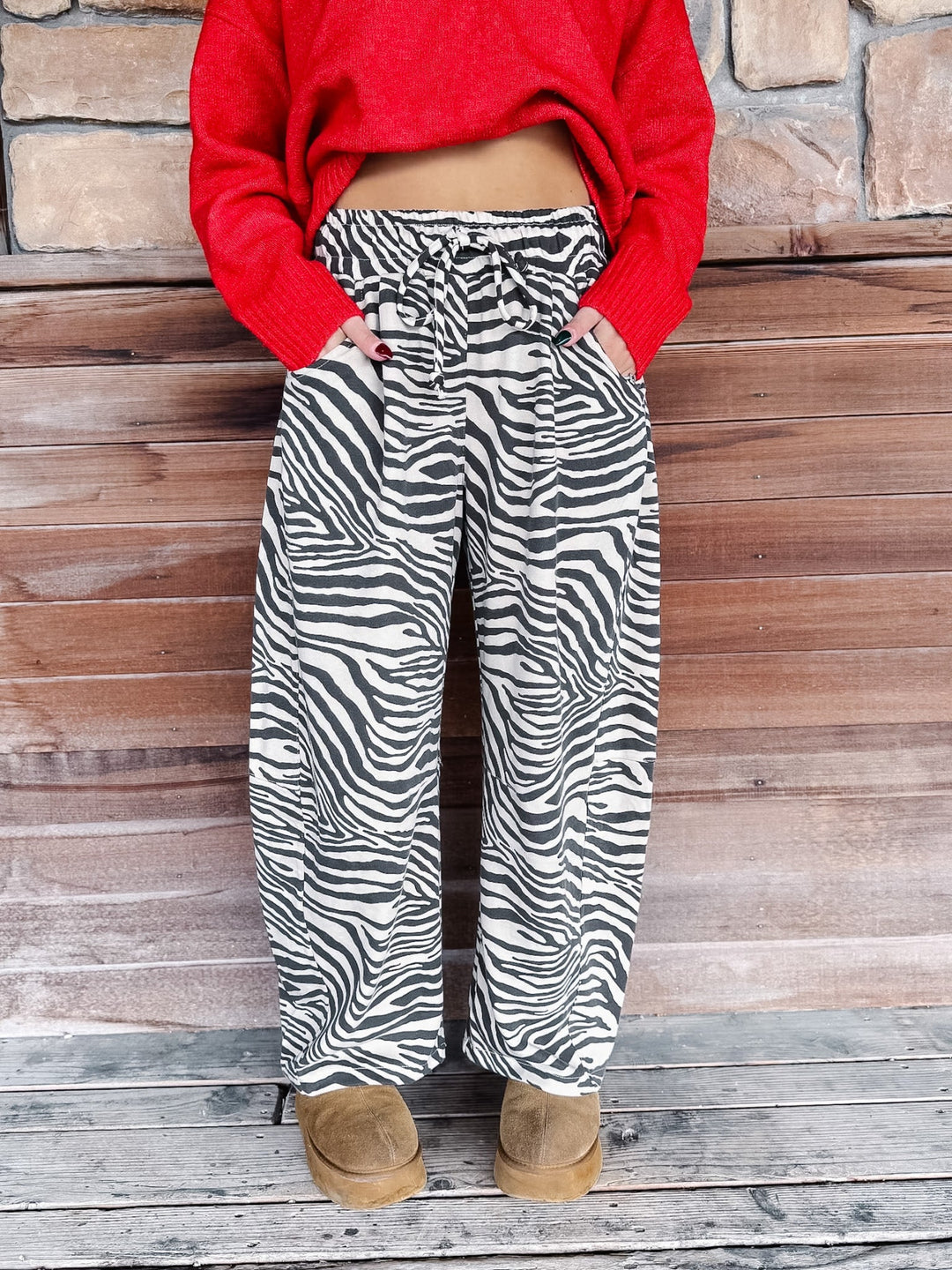 A person wearing black and white zebra print wide-leg pants, a bright red cropped sweater, and tan boots, standing against a rustic wooden and stone wall.
