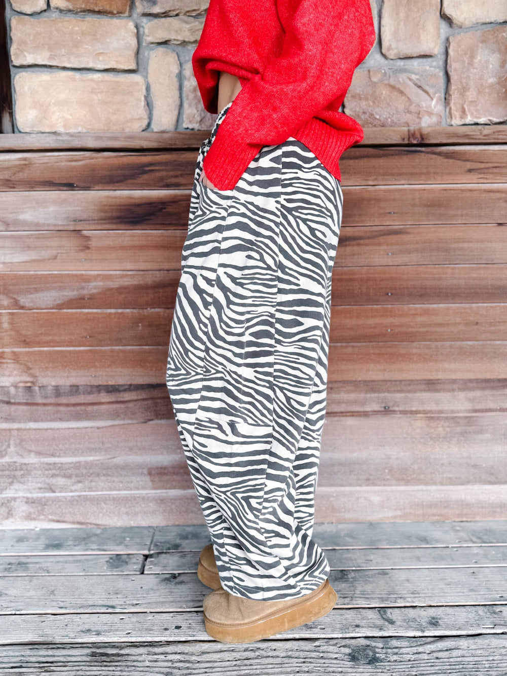 A person wearing loose black and white zebra-print pants, a bright red knit sweater draped off the shoulders, and tan slippers, standing against a rustic wooden wall and stone background.