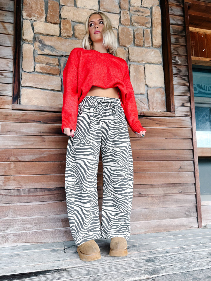 A woman stands on a wooden porch in front of a stone and wood wall, wearing a bright red cropped long-sleeve sweater and black-and-white zebra print wide-leg pants with tan boots.