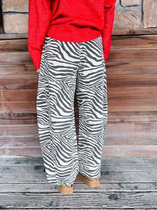 A person wearing wide-leg zebra-print pants and a bright red sweater, standing against a rustic wooden and stone wall.