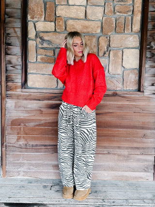 A woman stands on a rustic wooden porch in front of a stone wall, wearing a bright red knit sweater and black-and-white zebra print drawstring pants with tan boots.