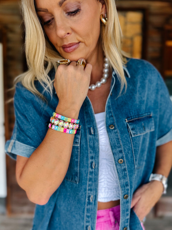A woman wearing a stack of colorful beaded bracelets with white pearl-like beads and gold-toned metal, styled with a white crop top, pink shorts, a pearl necklace, and a blue denim shirt.