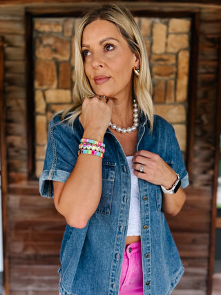 A woman wearing a blue denim short-sleeve button-up shirt over a white top and bright pink pants, accessorized with a pearl necklace and colorful bracelets, standing in front of a rustic stone and wood wall.