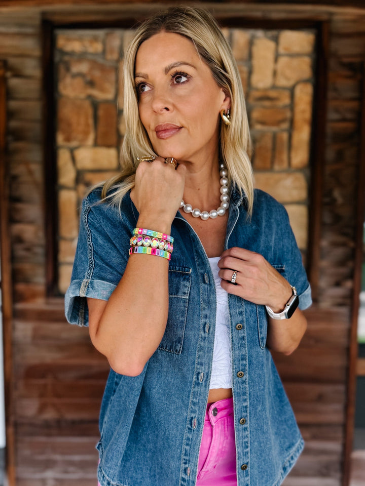 A woman wearing a blue denim short-sleeve button-up shirt over a white top and bright pink pants, accessorized with a pearl necklace and colorful bracelets, standing in front of a rustic stone and wood wall.